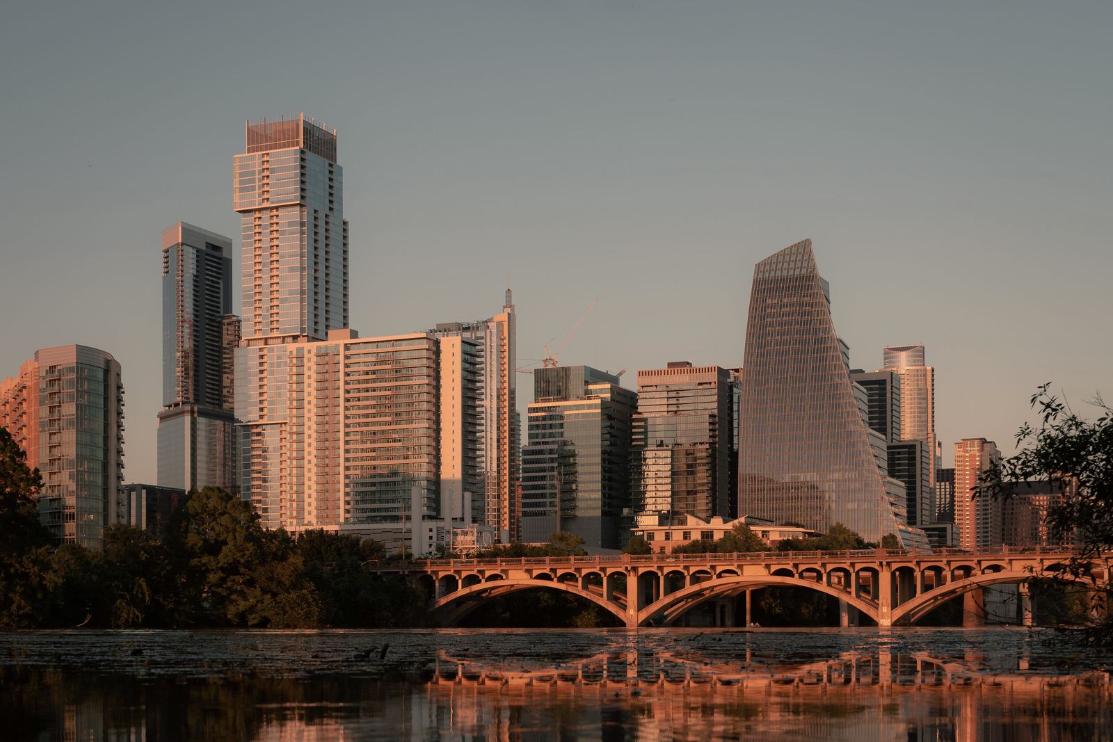 The Austin skyline at sunset, behind an arched bridge and reflected in still water.