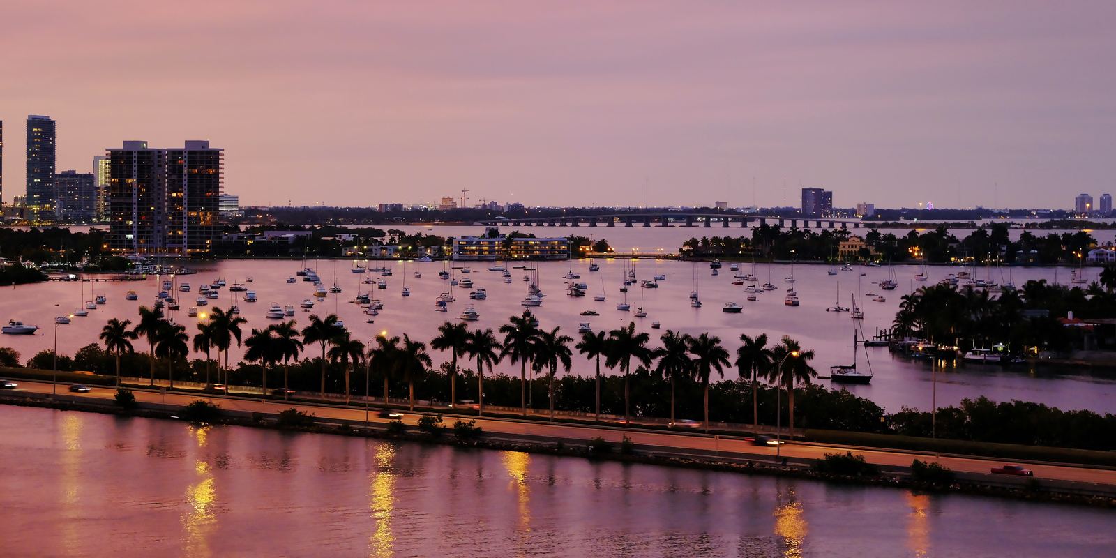 La bahía de Biscayne al anochecer, desde lo alto, con veleros fondeados, una hilera de palmeras y una calzada iluminada junto al agua.