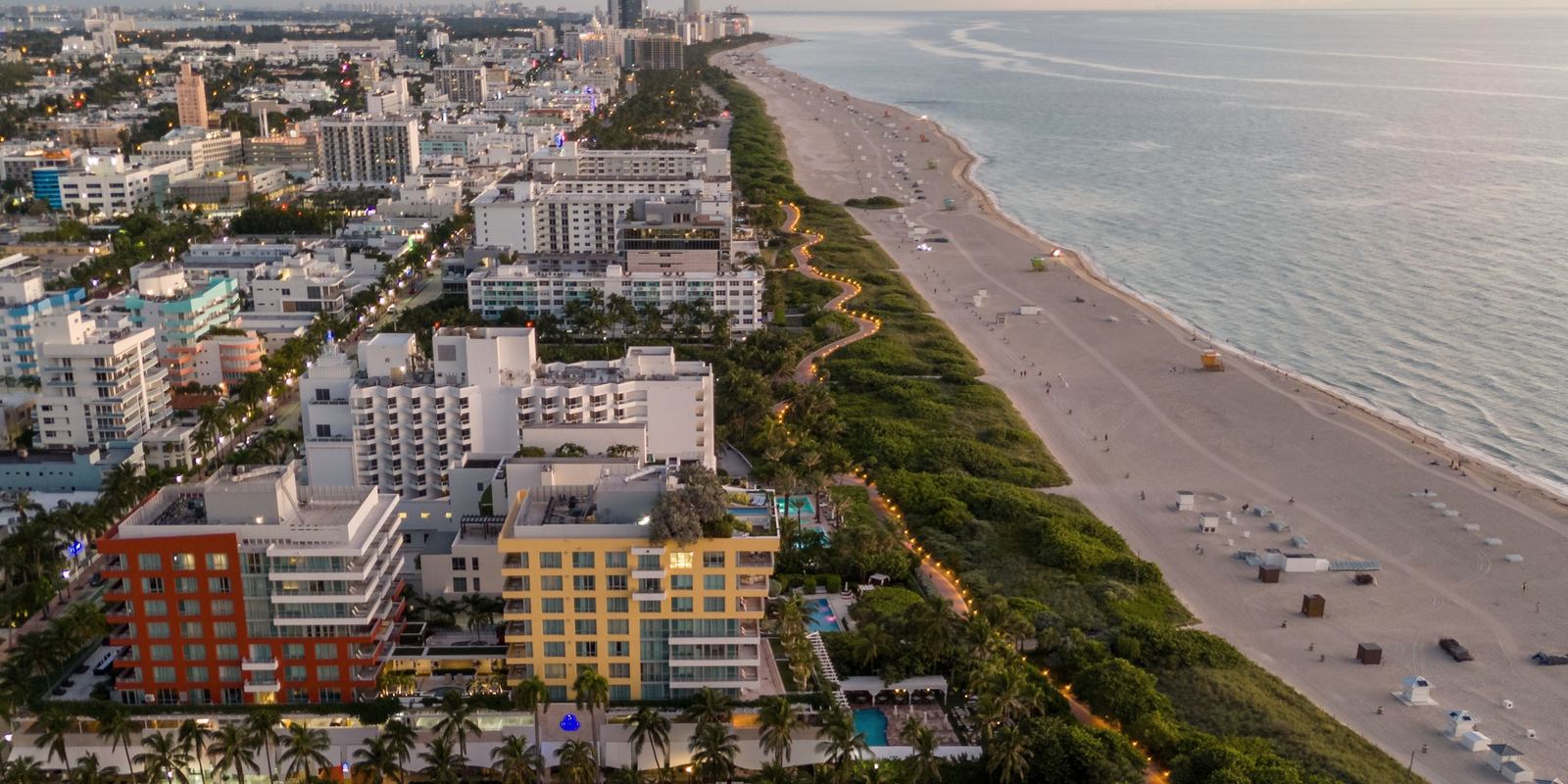 Miami Beach desde el aire al anochecer: hoteles y edificios bajos, un sendero iluminado en las dunas, una playa ancha y el Atlántico.