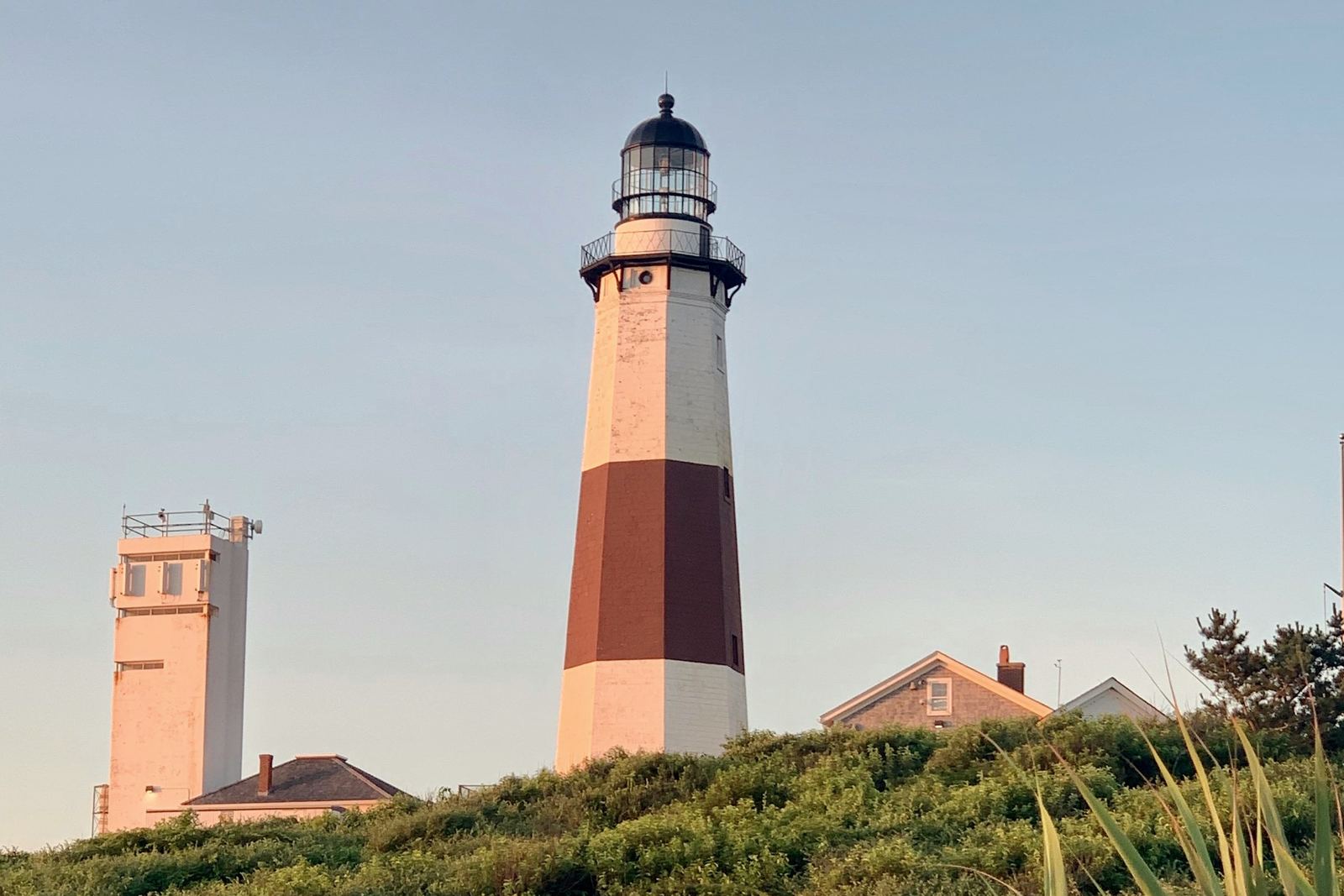 The striped Montauk Point Lighthouse on a green bluff in evening light, with beach grass in the foreground.