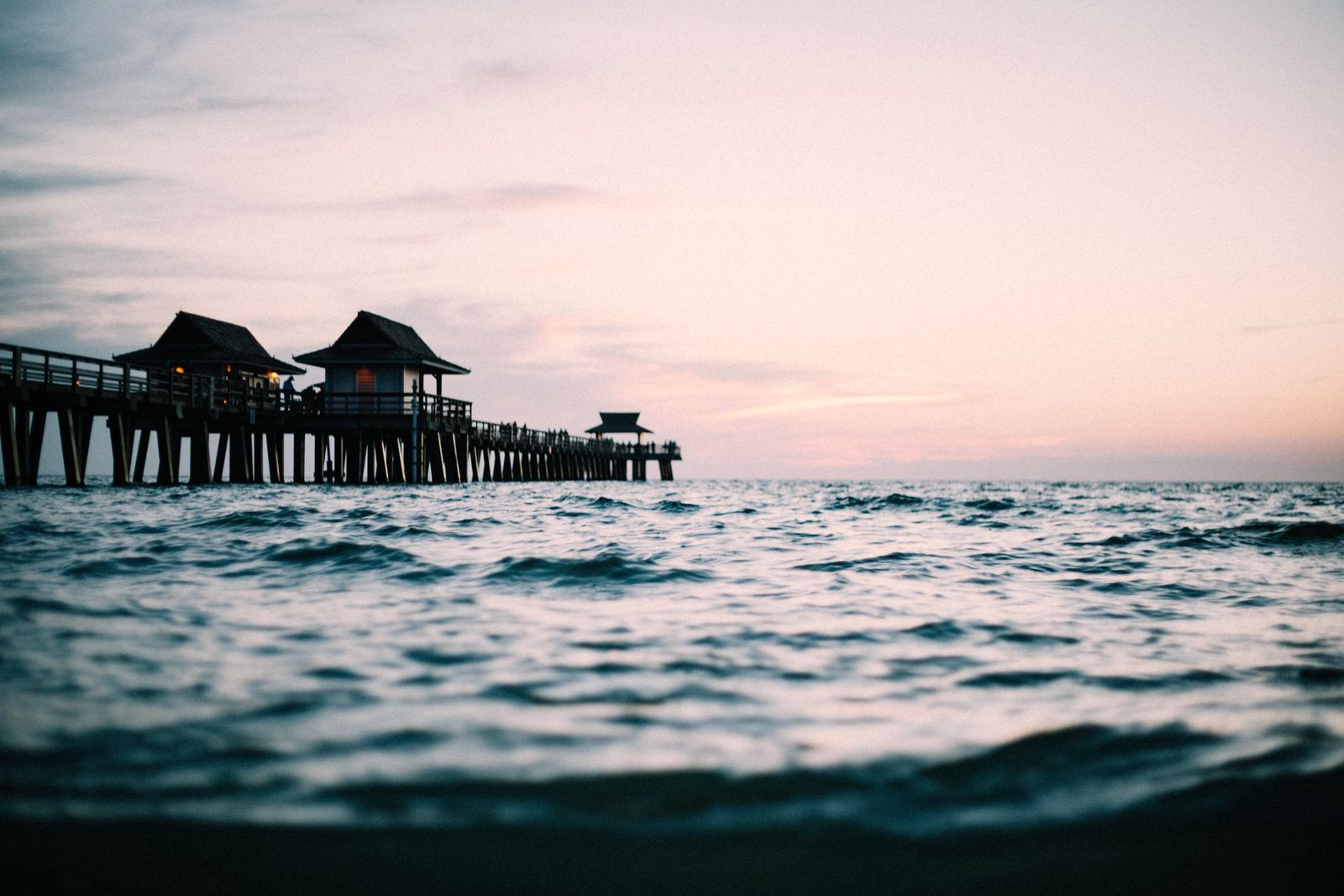 The Naples Pier at dusk under a pale pink sky, seen from the water.