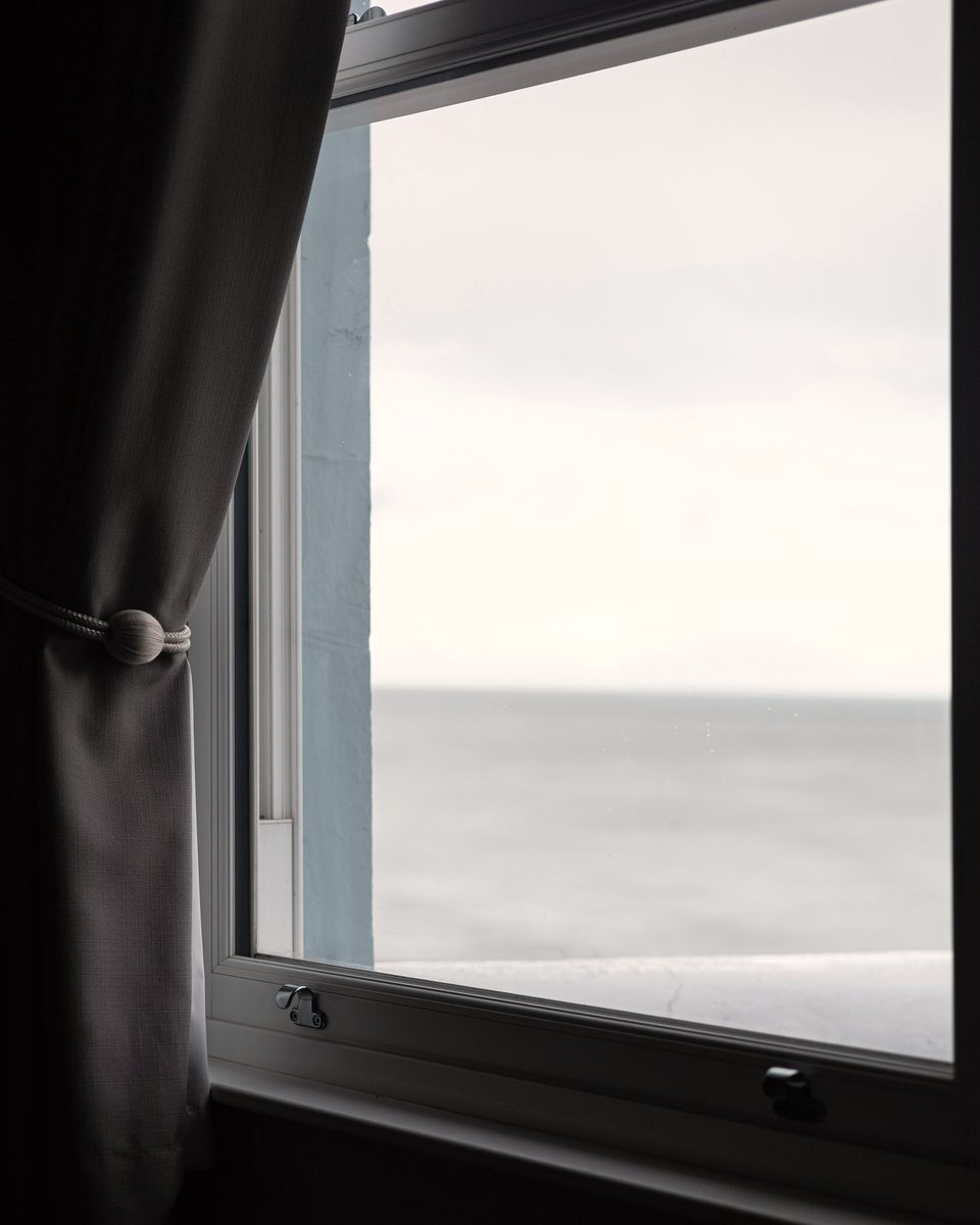 Un mar gris y en calma visto a través de una ventana abierta, con una cortina oscura recogida.
