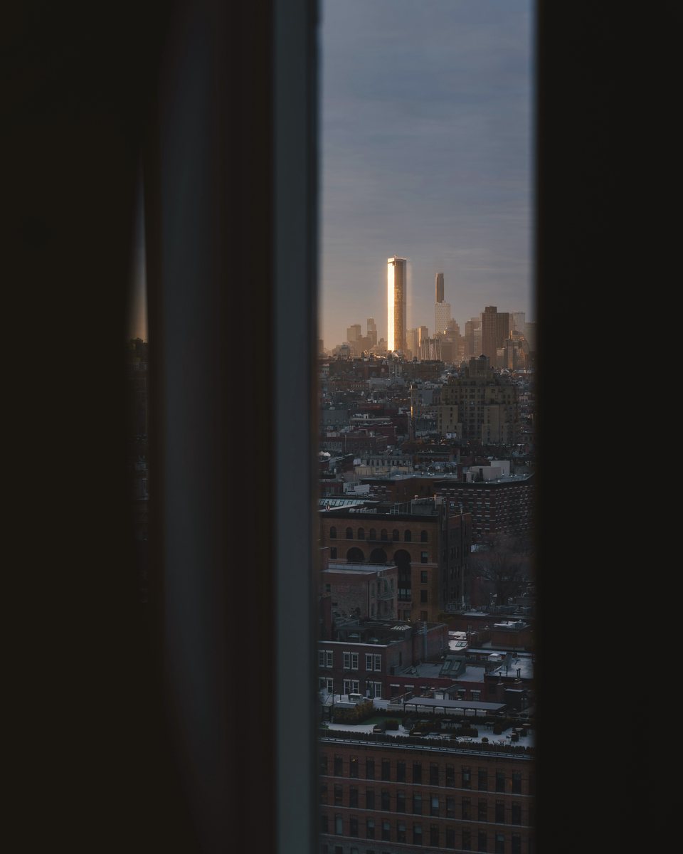 Torres de Manhattan iluminadas por el sol del atardecer, vistas por una ventana estrecha desde una habitación a oscuras.